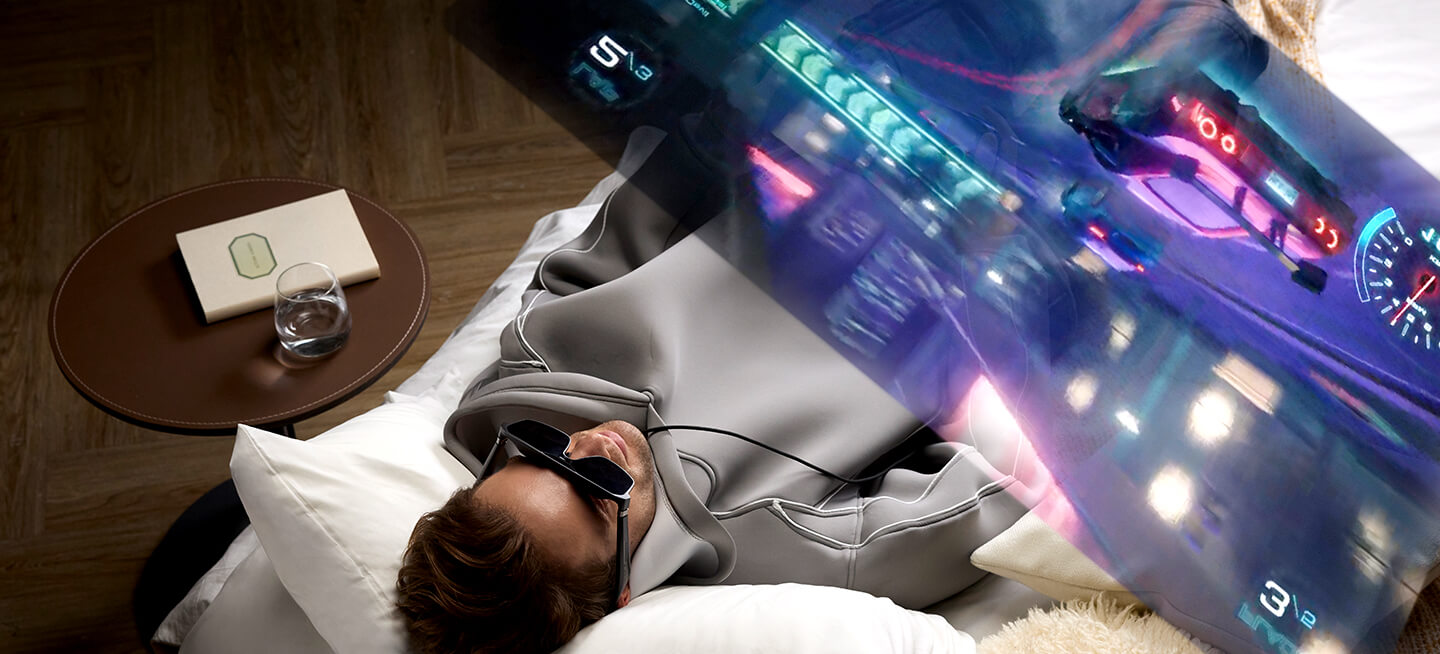 Мужчина лежит на кровати и наслаждается захватывающей игрой в очках ROG XREAL R1 и на консоли ROG Xbox Ally.